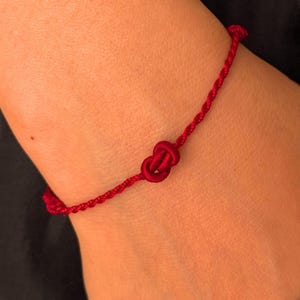 May include: A vibrant red string bracelet featuring a single central decorative knot. The band is constructed from a thin, twisted cord material that wraps around the wrist, secured by a small knot detail at the back of the bracelet design.
