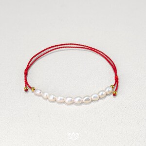 Peut inclure: Un bracelet en ficelle rouge orn&eacute; d'une rang&eacute;e de perles blanches de forme irr&eacute;guli&egrave;re. Le bracelet comporte des perles dor&eacute;es &agrave; chaque extr&eacute;mit&eacute;. Les perles sont enfil&eacute;es sur un fin cordon rouge.