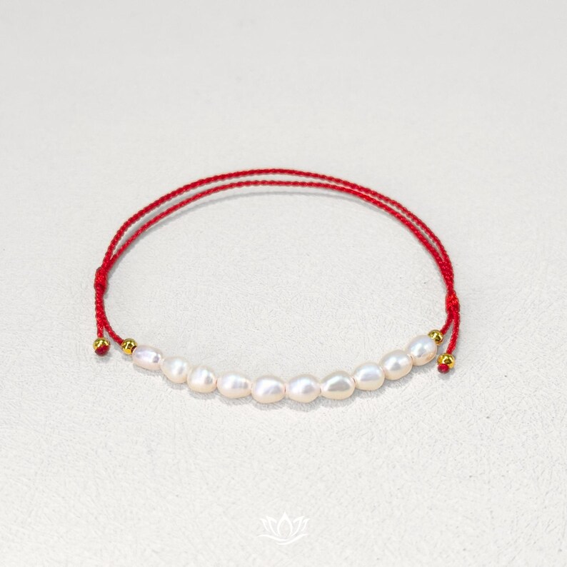 Peut inclure: Un bracelet en ficelle rouge orn&eacute; d'une rang&eacute;e de perles blanches de forme irr&eacute;guli&egrave;re. Le bracelet comporte des perles dor&eacute;es &agrave; chaque extr&eacute;mit&eacute;. Les perles sont enfil&eacute;es sur un fin cordon rouge.