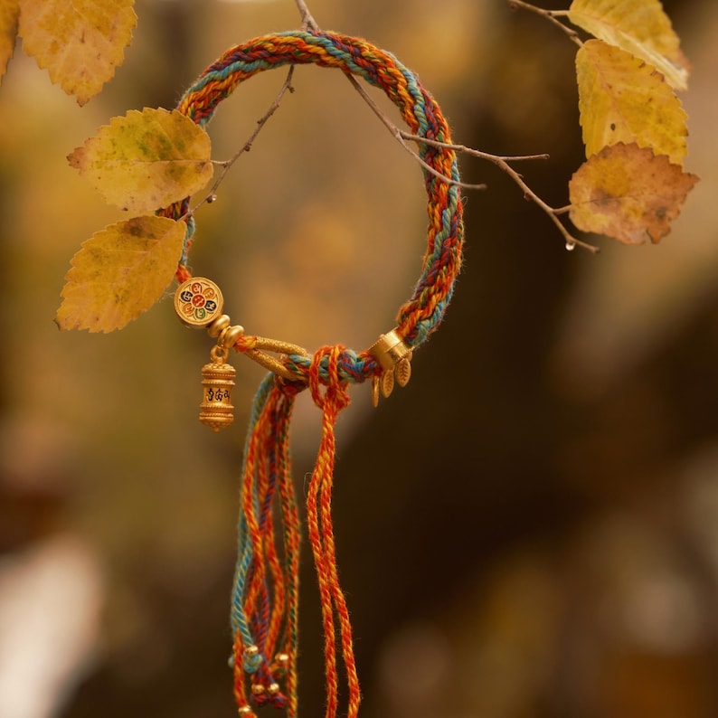 Pu&ograve; includere: Un braccialetto intrecciato colorato con dettagli dorati e ciondoli pendenti. Il braccialetto presenta fili blu, arancioni e verdi. Lo sfondo include foglie gialle e un ambiente naturale sfocato.