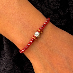 May include: Red and silver braided bracelet with a silver hexagonal charm. The charm has the number 69 on it. The bracelet is worn on a wrist.