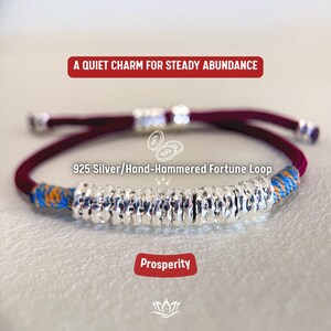 May include: A burgundy and silver bracelet with the text "925 Silver/Hand-Hammered Fortune Loop." The bracelet features a silver central section and blue and orange accents. The text "A Quiet Charm for Steady Abundance" and "Prosperity" are also present.