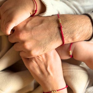 Puede incluir: Varias muñecas lucen pulseras de hilo rojo adornadas con cuentas doradas. Las pulseras presentan un fino cordón rojo y pequeñas cuentas doradas redondas. La imagen sugiere un tema de conexión o sentimiento compartido, con un diseño minimalista.