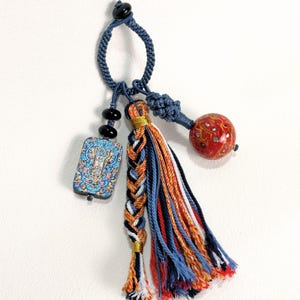 Könnte beinhalten: Ein dekorativer Schlüsselanhänger mit einer blauen geflochtenen Kordelschlaufe, einem rechteckigen Anhänger mit farbenfrohem Design und einer roten Kugel mit Muster. Der Schlüsselanhänger enthält auch eine geflochtene Quaste mit mehreren Farben.