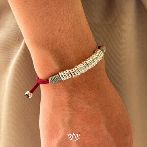 May include: Silver bracelet featuring a series of small, textured silver rings. The bracelet has a red, blue, and yellow woven cord. The bracelet is adjustable with a sliding knot closure. The bracelet is shown on a person's wrist.