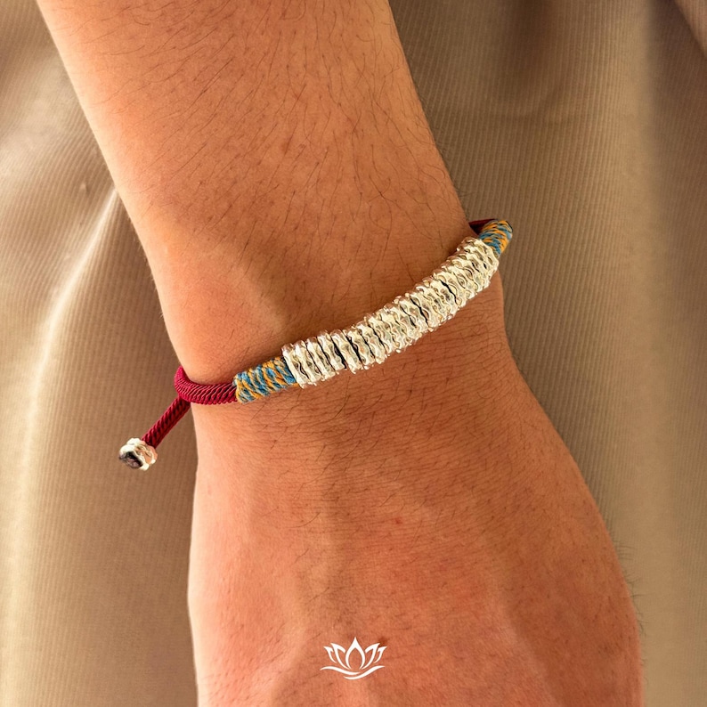 May include: Silver bracelet featuring a series of small, textured silver rings. The bracelet has a red, blue, and yellow woven cord. The bracelet is adjustable with a sliding knot closure. The bracelet is shown on a person's wrist.