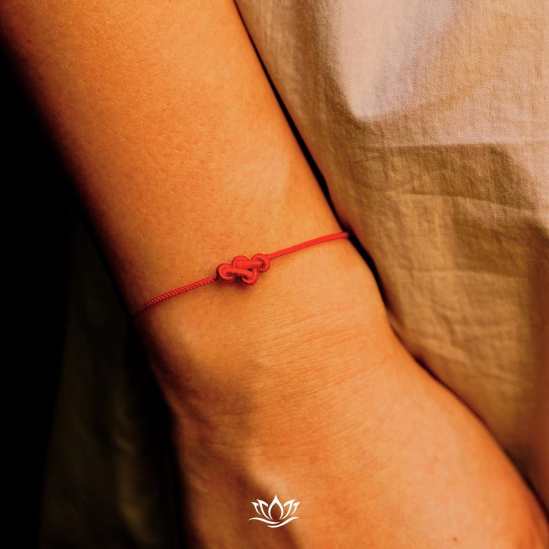 Pode incluir: Uma pulseira de fio vermelho com um n&oacute; decorativo no centro. A pulseira &eacute; usada no pulso, com o fio vermelho contrastando com o tom de pele. O fundo &eacute; de uma cor suave e neutra.