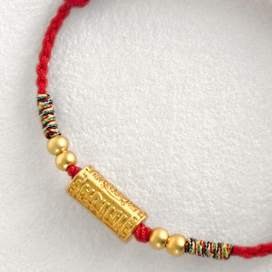 May include: A red braided cord bracelet featuring a central gold-colored cylindrical bead with embossed geometric patterns and text, flanked by two small gold-colored spherical beads on each side and multicolored thread accents.