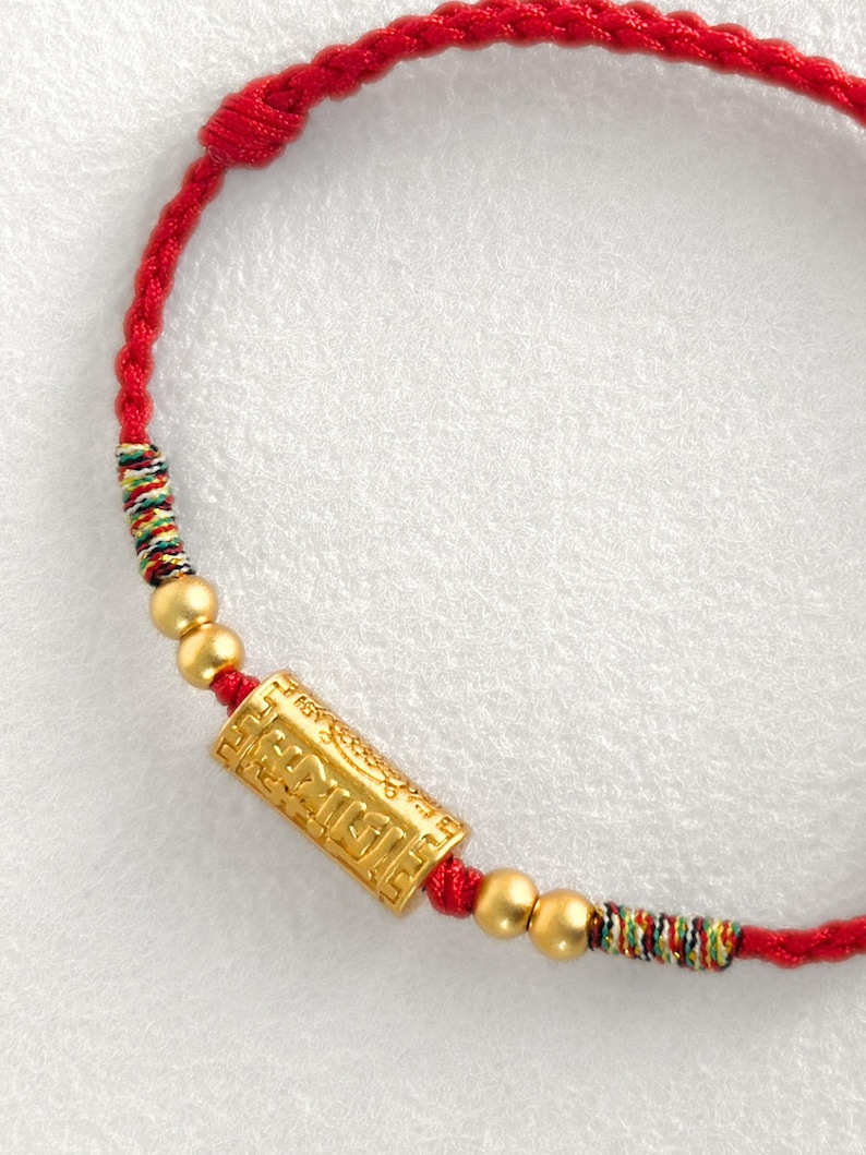 May include: A red braided cord bracelet featuring a central gold-colored cylindrical bead with embossed geometric patterns and text, flanked by two small gold-colored spherical beads on each side and multicolored thread accents.