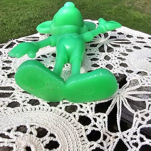 Puede incluir: Una figura verde brillante, de estilo vintage, de un personaje de dibujos animados, tumbada boca arriba con los brazos y las piernas extendidos. La figura descansa sobre un mantel de ganchillo blanco, con c&eacute;sped verde y &aacute;rboles al fondo.