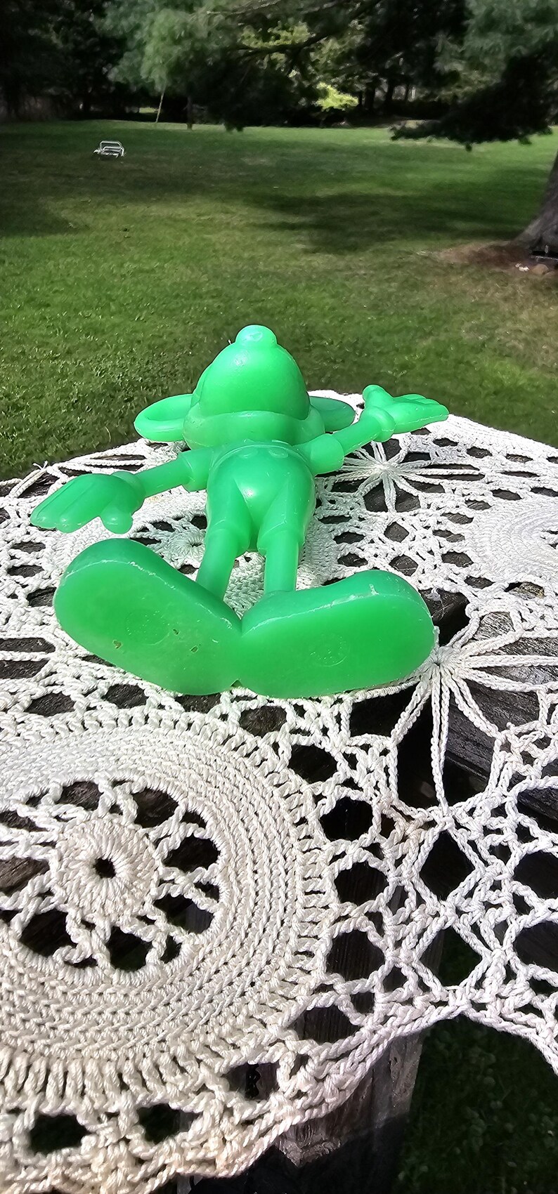 Puede incluir: Una figura verde brillante, de estilo vintage, de un personaje de dibujos animados, tumbada boca arriba con los brazos y las piernas extendidos. La figura descansa sobre un mantel de ganchillo blanco, con c&eacute;sped verde y &aacute;rboles al fondo.
