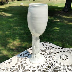 May include: A white ceramic goblet with a smooth, rounded bowl and a textured, tree-trunk-inspired stem that flares out at the base. The surface of the vessel has a glossy finish, reflecting light across its curved form and detailed, organic stem structure.
