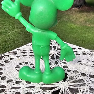 Puede incluir: Una figura verde de Mickey Mouse con el brazo extendido y orejas grandes. La figura est&aacute; sobre un mantel individual blanco de ganchillo. El fondo presenta una zona de c&eacute;sped y &aacute;rboles.