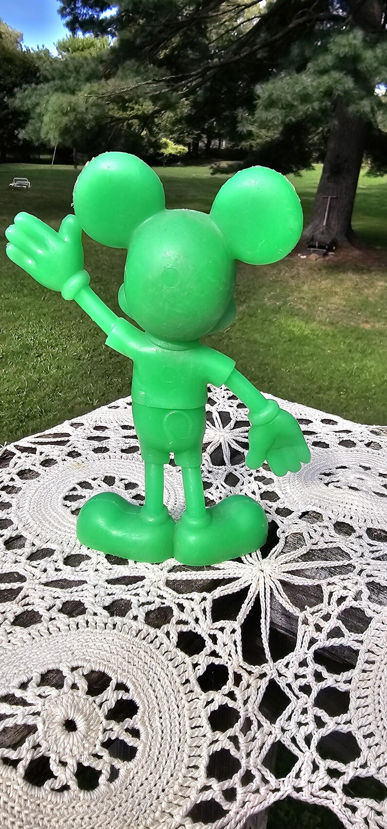 Puede incluir: Una figura verde de Mickey Mouse con el brazo extendido y orejas grandes. La figura est&aacute; sobre un mantel individual blanco de ganchillo. El fondo presenta una zona de c&eacute;sped y &aacute;rboles.