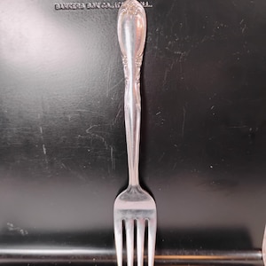 May include: A silver fork with an ornate handle, set against a dark background. The fork has four tines and a detailed design on the handle. The text "COPYHOLDER NO.76 BANKERS BOX CO., IL" is visible in the background.