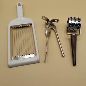 May include: A white egg slicer, a silver can opener, and a brown-handled meat tenderizer are arranged on a light tan surface. The egg slicer has a rectangular frame with parallel wires. The can opener is made of silver metal. The meat tenderizer has a roller with spikes.