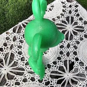 Puede incluir: Una figura de delf&iacute;n de pl&aacute;stico verde brillante con una aleta en forma de cactus, apoyada sobre un tapete de ganchillo blanco. El delf&iacute;n es de color s&oacute;lido y parece ser un juguete o decoraci&oacute;n vintage. El fondo es una superficie de madera y hierba.