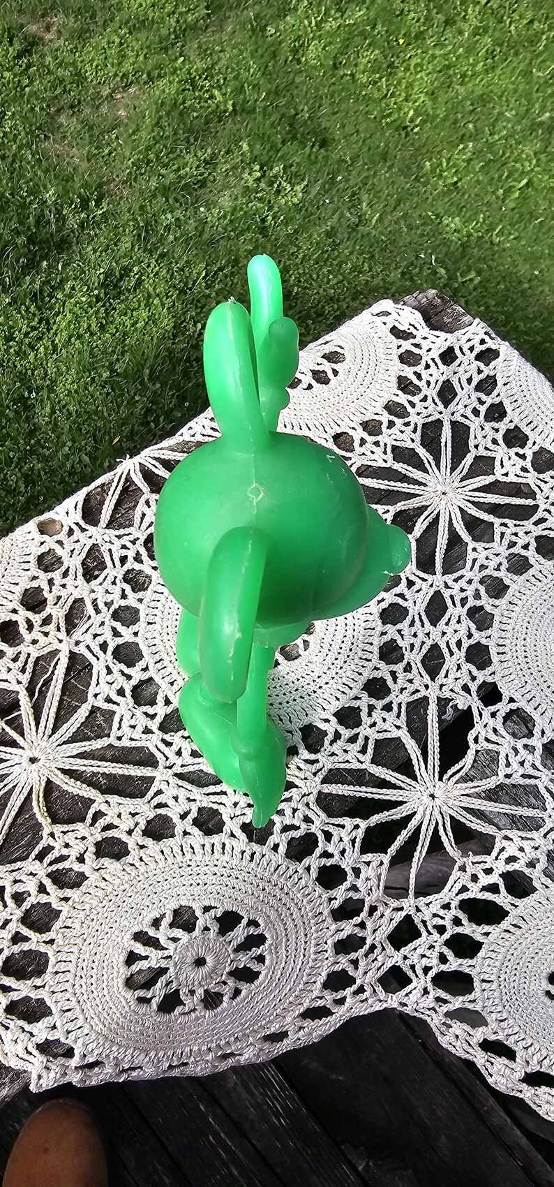 Puede incluir: Una figura de delf&iacute;n de pl&aacute;stico verde brillante con una aleta en forma de cactus, apoyada sobre un tapete de ganchillo blanco. El delf&iacute;n es de color s&oacute;lido y parece ser un juguete o decoraci&oacute;n vintage. El fondo es una superficie de madera y hierba.