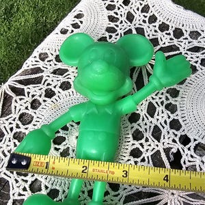 Puede incluir: Una figura de Mickey Mouse de pl&aacute;stico verde, de aproximadamente 10 cm de altura, con los brazos extendidos. Una cinta m&eacute;trica amarilla Stanley se coloca cerca de la figura. El fondo incluye un mantelito de encaje blanco y hierba verde.