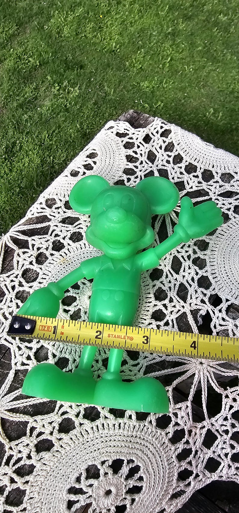 Puede incluir: Una figura de Mickey Mouse de pl&aacute;stico verde, de aproximadamente 10 cm de altura, con los brazos extendidos. Una cinta m&eacute;trica amarilla Stanley se coloca cerca de la figura. El fondo incluye un mantelito de encaje blanco y hierba verde.