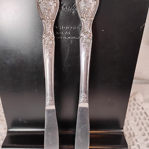 May include: Two silver butter knives with ornate floral and scrollwork handles. The knives are displayed against a black background, with the text "Butterholder No. 76" visible. The knives are likely vintage or antique.