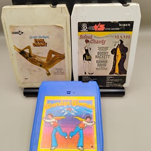 May include: Three vintage 8-track tapes. The top two are off-white with album art and text. The bottom tape is blue with a colorful image of two figures and the text "Wonder Women".