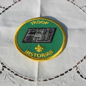 May include: A green and gold embroidered patch with the text "TROOP HISTORIAN" and a black and white illustration of an open book.