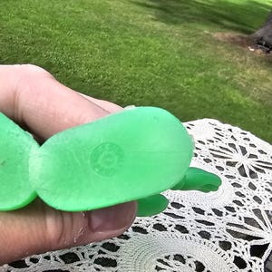 Puede incluir: Un juguete de pl&aacute;stico verde brillante, posiblemente una tortuga o criatura similar, se sostiene en una mano. El juguete tiene una superficie texturizada y una marca circular en su parte inferior. El fondo muestra un c&eacute;sped verde y &aacute;rboles.