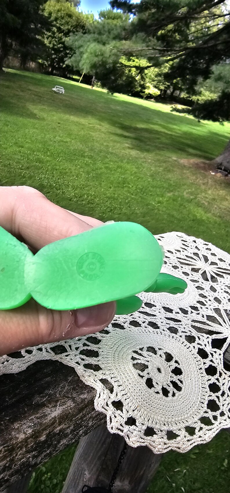 Puede incluir: Un juguete de pl&aacute;stico verde brillante, posiblemente una tortuga o criatura similar, se sostiene en una mano. El juguete tiene una superficie texturizada y una marca circular en su parte inferior. El fondo muestra un c&eacute;sped verde y &aacute;rboles.