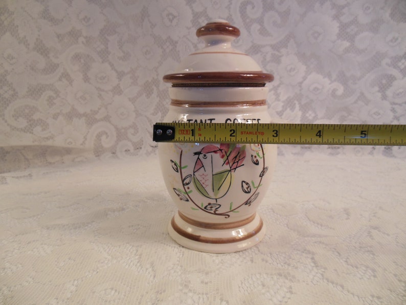 Peut inclure: Pot &agrave; caf&eacute; instantan&eacute; en c&eacute;ramique, d'environ 13 cm de haut, avec couvercle. Le pot est blanc cass&eacute; avec des accents bruns et un motif peint &agrave; la main d'oiseau et de fleurs. Le texte "INSTANT COFFEE" est imprim&eacute;.