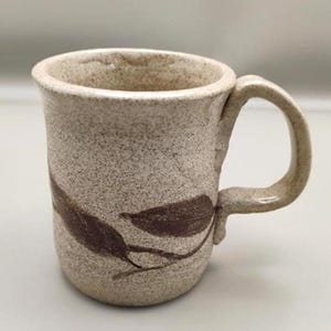 May include: A speckled beige ceramic mug with a textured, grainy surface. The mug features a dark brown botanical leaf design wrapped around the lower exterior and includes a sturdy, curved handle attached to the side of the cylindrical body.