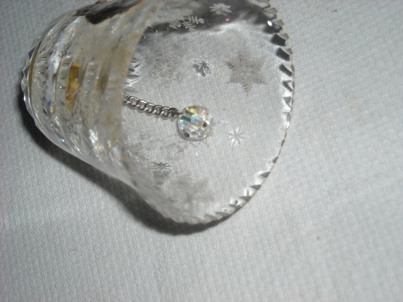 K&ouml;nnte beinhalten: Eine klare Glasglocke mit einem Kristall, der an einer Kette im Inneren h&auml;ngt. Die Glocke hat ein dekoratives, ge&auml;tztes Muster aus Schneeflocken.