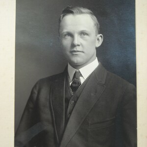 May include: A black and white portrait photograph of a man wearing a suit and tie. He is looking directly at the camera with a serious expression.