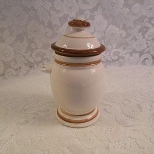 Peut inclure: Un pot en c&eacute;ramique blanc avec un couvercle, orn&eacute; de d&eacute;tails marron et dor&eacute;s. Le pot a un corps arrondi, une base en pi&eacute;destal et de petites poign&eacute;es. Le couvercle est surmont&eacute; d'un petit bouton marron. Le pot est probablement pour le stockage ou l'affichage.