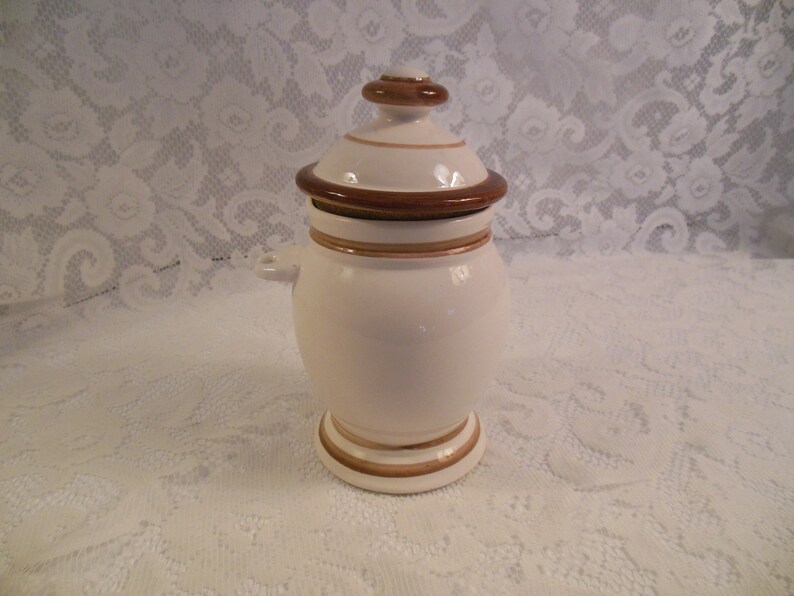 Peut inclure: Un pot en c&eacute;ramique blanc avec un couvercle, orn&eacute; de d&eacute;tails marron et dor&eacute;s. Le pot a un corps arrondi, une base en pi&eacute;destal et de petites poign&eacute;es. Le couvercle est surmont&eacute; d'un petit bouton marron. Le pot est probablement pour le stockage ou l'affichage.