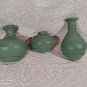 May include: Three green decorative vases of varying sizes. The vases have a matte finish and a classic shape, with a rounded body and a narrow neck. The vases are arranged on a white surface.