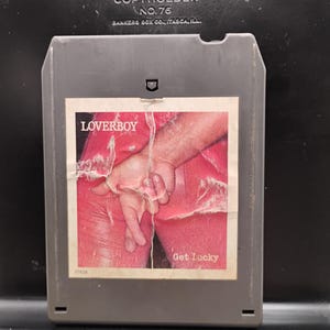 May include: A vintage Loverboy 8-track tape. The label features the band's name and the title "Get Lucky." The image on the label shows a person's crossed fingers. The tape is in a gray plastic cartridge.