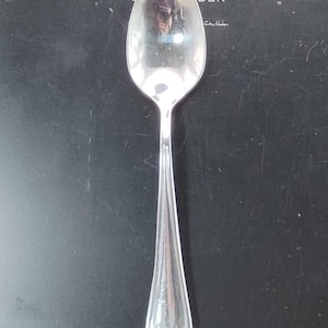 May include: A silver spoon with a long, slender handle and an oval bowl. The handle has a decorative floral design at the base. The spoon reflects the surrounding environment.
