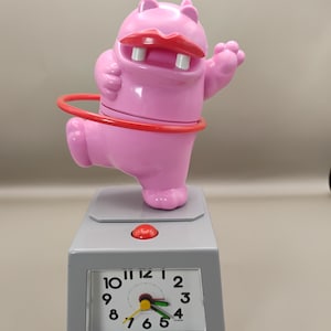 May include: A pink hippo-shaped alarm clock with a red hula hoop and red lips. The clock has a gray base with a red button and a clock face showing the time. The text "shot on moto g 5G - 2024 sweetcindylouwho" is visible.