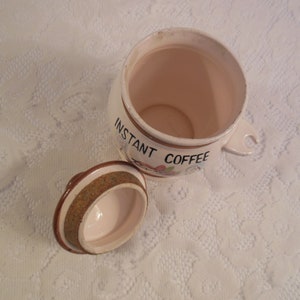 Peut inclure: Un pot en c&eacute;ramique blanc cass&eacute; ouvert avec un couvercle, con&ccedil;u pour le caf&eacute; instantan&eacute;. Le pot a une poign&eacute;e et est d&eacute;cor&eacute; des mots "INSTANT COFFEE" et d'&eacute;l&eacute;ments floraux. Le couvercle est sur le c&ocirc;t&eacute;.