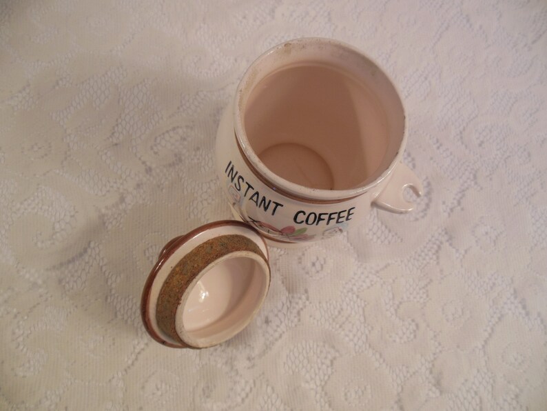 Peut inclure: Un pot en c&eacute;ramique blanc cass&eacute; ouvert avec un couvercle, con&ccedil;u pour le caf&eacute; instantan&eacute;. Le pot a une poign&eacute;e et est d&eacute;cor&eacute; des mots "INSTANT COFFEE" et d'&eacute;l&eacute;ments floraux. Le couvercle est sur le c&ocirc;t&eacute;.
