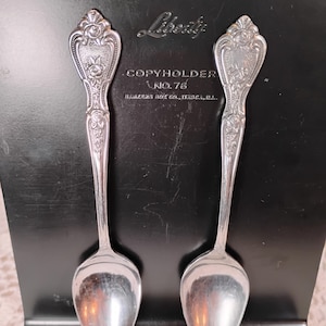 May include: Two silver spoons with ornate floral designs on the handles, resting against a black copyholder. The copyholder has the word "Liberty" and the text "COPYHOLDER NO. 76" embossed on it. The spoons are reflective.