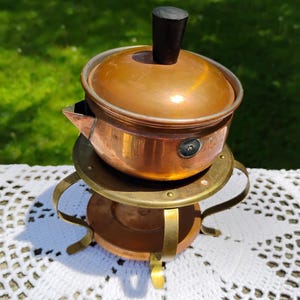 May include: A copper pot with a lid featuring a black knob, resting on a brass stand with three curved legs. The pot has a small spout and a circular side attachment. The metal surfaces show a warm, reflective finish and signs of age.