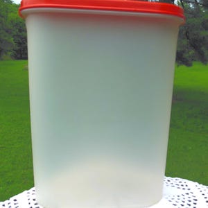 May include: A tall, rectangular, frosted plastic container with a bright red lid. The container is sitting on a white, lace-like surface, with a green grassy background.
