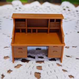 May include: A miniature brown plastic roll-top desk featuring a flat work surface, a hutch with two small side compartments, and two pedestals with molded drawer fronts. The desk has a smooth finish and a classic structure with a top shelf.
