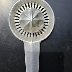 May include: A vintage, silver-toned Foley citrus juicer. The juicer has a round, perforated head with a star-shaped center and a long handle with a hole for hanging. The word "Foley" is engraved on the handle.
