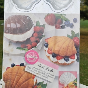 May include: A silver-colored, shell-shaped baking mold with two cavities, displayed with a box showing shell-shaped desserts. The box features the text "Mini Shells" and images of desserts with berries. The mold is designed for making elegant desserts and salads.