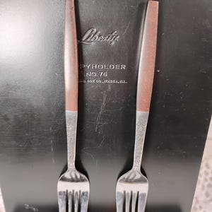 May include: Two vintage forks with brown handles and ornate silver tines. The forks are displayed against a black background with the word "Liberty" and "PYHOLDER NO. 76" visible. The forks are likely part of a vintage flatware set.