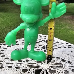 Puede incluir: Una figura verde de Mickey Mouse, de aproximadamente 18 cm de altura, sobre un tapete blanco de ganchillo. La figura tiene los brazos extendidos y orejas grandes, con una cinta m&eacute;trica amarilla en el fondo.