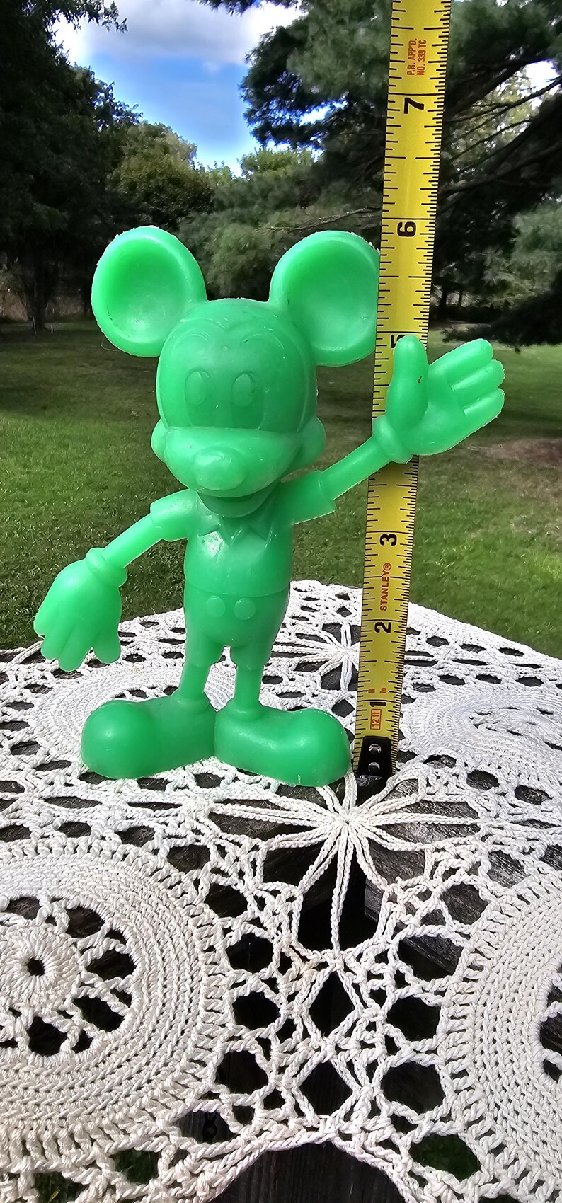 Puede incluir: Una figura verde de Mickey Mouse, de aproximadamente 18 cm de altura, sobre un tapete blanco de ganchillo. La figura tiene los brazos extendidos y orejas grandes, con una cinta m&eacute;trica amarilla en el fondo.